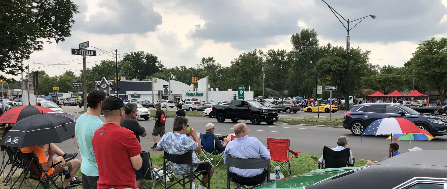 Spectators in lawn chairs and umbrellas on the grass at Woodward and Sheffield, watching cars roll by on the boulevard.